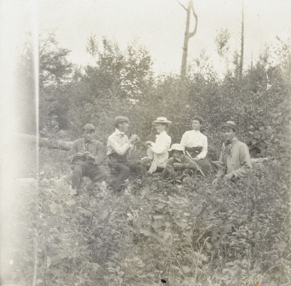 Looking at each other, Lucy and W.A. Holt are eating berries together. Bertha {Unknown} is sitting to the right of Lucy Holt. One boy is sitting on a fallen tree to the left of W.A. Holt. Another boy is crouched next to the tree, and a third boy is standing nearby. 