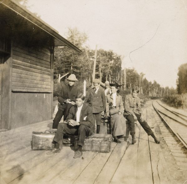View down platform of Lakewood train station, with the McClure family waiting to board a train. Names from left to right: Dr. James McClure, Jim McClure, Archibald McClure, Mrs. Annie McClure, and Nathan McClure. There are railroad tracks on the right.