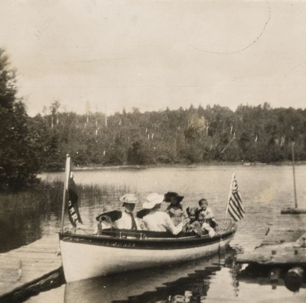 The <i>Islander</i> steam launch is docked at the pier on the north side of the Island. There are two women and some children in the picture. Jeannette, Eleanor, and Alfred Holt are in the back of the boat. The <i>Islander</i> flag is waving at the bow, and a flag with an anchor and stripes is flying at the stern of the boat. 