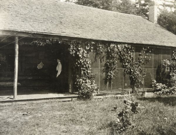 View of Island Lodge, with a front porch on the left, and a screened-in porch on the right. A woman, possibly Lucy Holt, is standing on the porch. 