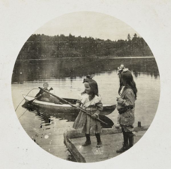 Wearing a dress, Donald Holt is fishing, using a wooden pole, in Archibald Lake. Alfred Holt is paddling a canoe. Eleanor Holt is standing next to Donald on the pier and watching Alfred. 