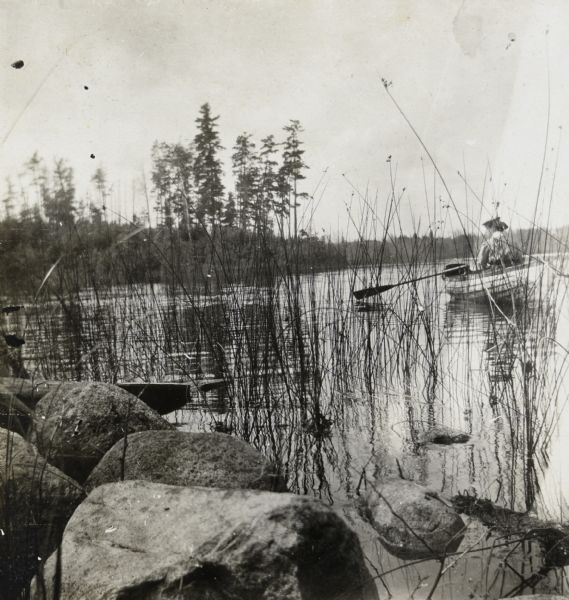 With her back to the camera, Emily McDonald is rowing a boat on Archibald Lake. Large rocks and grasses are in the foreground. Caption reads: "Emily McDonald - Aug. 3rd."