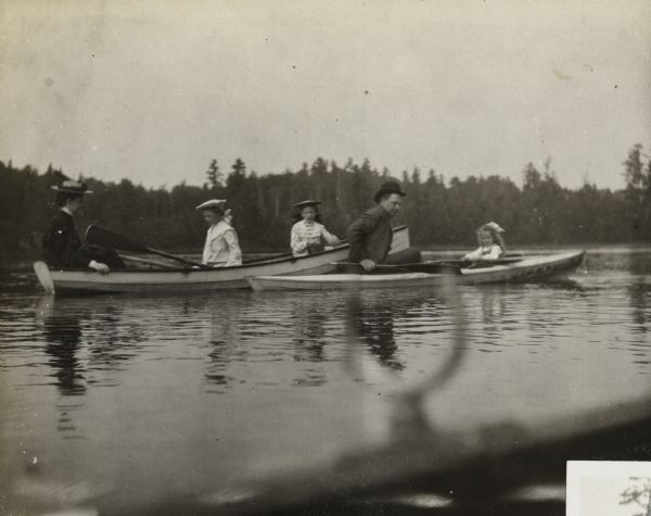 View from boat across water towards a group of five out in two boats on Archibald Lake. On the left, Harriet Stroh is rowing while Ellen Holt is sitting and watching her, and in the front of the boat is Jeannette Holt sitting and looking at the camera. In the clinker-built duck boat on the right are Mr. Delbridge and Eleanor Holt, both holding paddles. The tree-lined shore is in the far background. Caption reads: "Ellen Holt, Mr. Delbridge." 