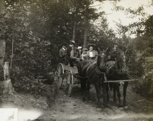 Mr. and Mrs. Delbridge, with a group of people, are riding in a horse-drawn wagon on a dirt road. The two horses are wearing leather bridles. Caption reads: "Mr. and Mrs. Delbridge - Aug 5th."