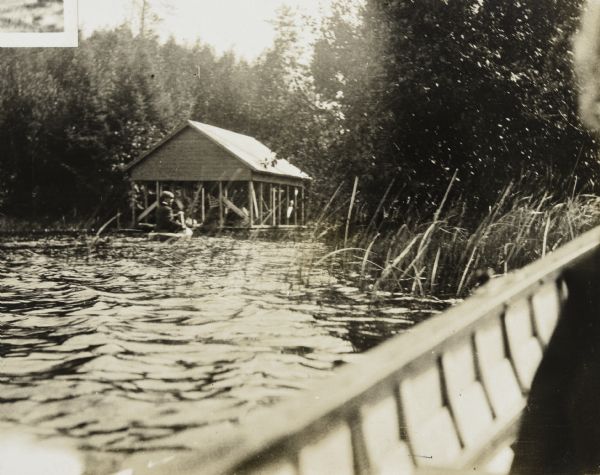 View across water from a boat on Archibald Lake towards a man paddling a duck boat toward the new boathouse on the island. The <i>Islander </i>steam launch, with a nautical flag flying from the stern, is parked in the boathouse. Trees and grasses are growing along the shoreline. Caption reads: "The new boat house."