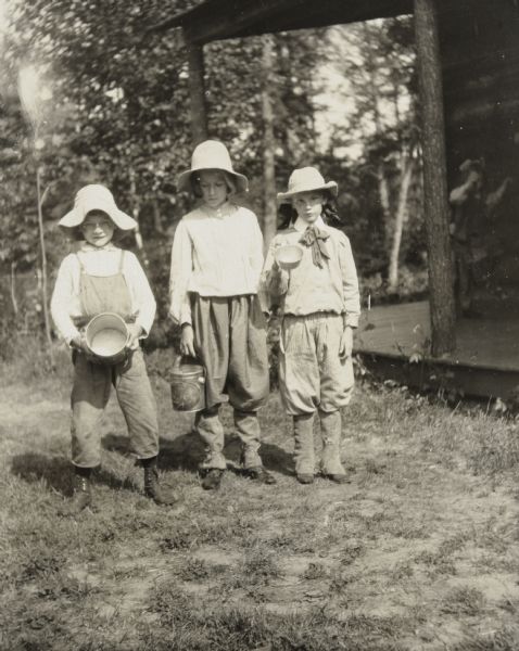 Three children, wearing sun hats, are holding empty berry containers. The girls are wearing knickers. On the right is the Island Lodge porch. From left to right are: Alfred Holt, Harriet Stroh, and Jeannette Holt. Caption reads: "Off for berries. Alfred, Harriet, and Jeannette." 