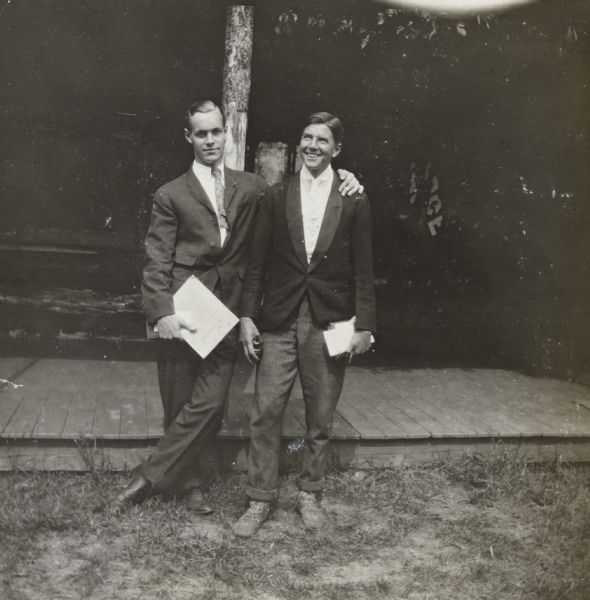 Standing in front of the Island Lodge porch, two young men are dressed for church on Sunday. On the left, John Stevenson is wearing a suit and tie and holding a book and papers. He is resting his hand on James McClure's shoulder. On the right, McClure is wearing a tuxedo jacket, vest and tie and holding a book and papers. Caption reads: "Disguised for Sunday — John Stevenson and James."