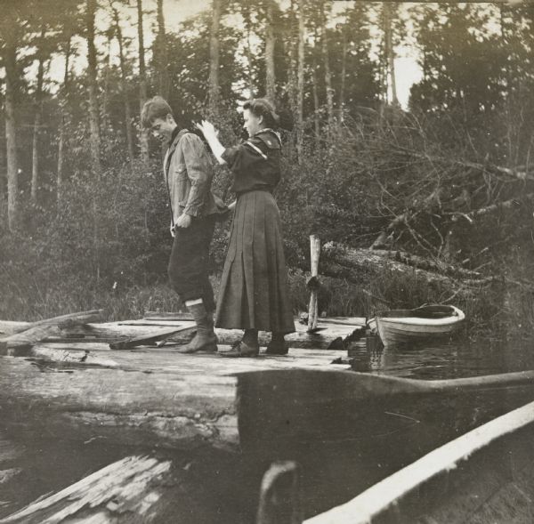 View from boat at Archibald Lake towards Isabell Holt (?) and Joe Rumsey (?) standing on the pier. Holt appears to be brushing off the back of Rumsey's jacket. There is a boat in the background which has been pulled up on the shore where trees and grasses are growing. Caption reads: "A New Cleaning Process — Isabell and Joe."