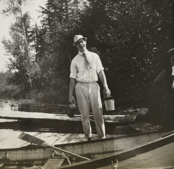 A man is holding two small metal pails. A woman, just out of frame, is standing on the far right. A rowboat is in the foreground, and another pier is in the background. The shoreline of Picnic Island with trees and grasses is in the background on the right.


