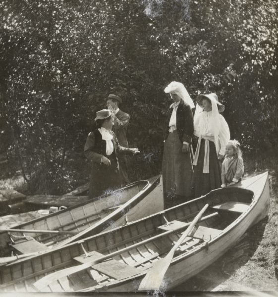 View towards shoreline of Archibald Lake where a group of five people, three women, one man, and one child, is getting ready for a picnic. Two rowboats are pulled up on shore. Trees are in the background. Photo album page caption reads: "Setting off for a picnic." 