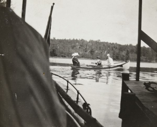 View from the new boathouse on the Island towards a foursome out boating on Archibald Lake. A man is rowing, and one woman, sitting on a bench in the middle of the boat, is looking toward the back where two other women, wearing hats, are sitting. The <i>Islander</i> steam launch, parked in the new boathouse, is in the foreground.