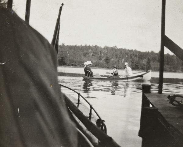 View from the new boathouse on the Island towards a foursome out boating on Archibald Lake. A man is rowing, and one woman, sitting on a bench in the middle of the boat, is looking toward the back where two other women, wearing hats, are sitting. The <i>Islander</i> steam launch, parked in the new boathouse, is in the foreground. 