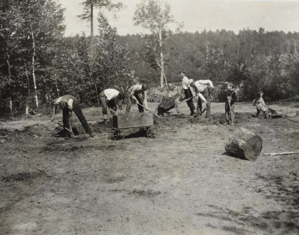 A group of five men are preparing the soil for the new tennis court. The men are using shovels, rakes, and wheelbarrows. On the right, children Alfred and Donald Holt are helping to build the tennis court. A tree stump is lying in the foreground. Archibald Lake and a forest on the mainland are in the background. Caption reads: "The tennis court crew." 


