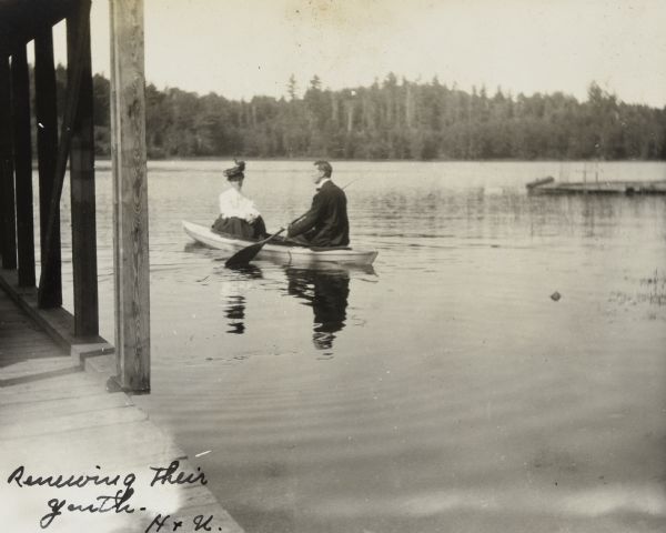 View from the new boathouse towards a couple canoeing on Archibald Lake. The woman is looking at the camera. The man, in profile, is using a paddle. Across the lake is a forest. Caption reads: "Renewing their Youth. H & U." 