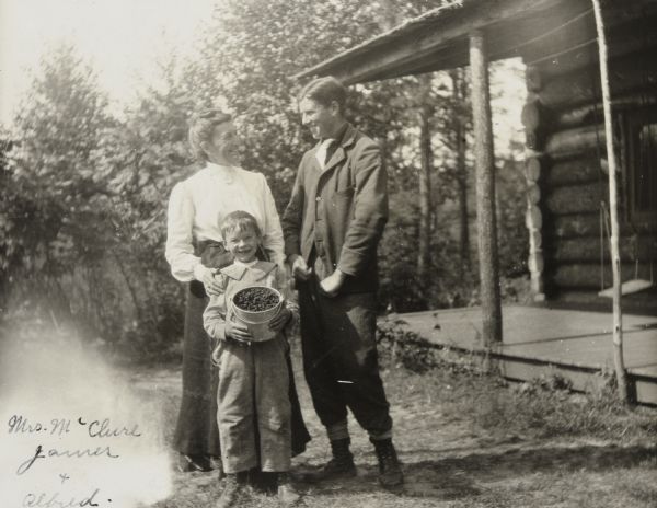 Looking at the camera, Alfred Holt, smiling, with a tooth missing, is holding a pail full of freshly-picked berries. Mrs. Annie McClure is standing behind Alfred and has her hand on his shoulder. Mrs. McClure and her son James McClure, standing on the right, are looking at each other and smiling. The Island Lodge porch, with a swing, is on the far right. Trees are in the background. 

