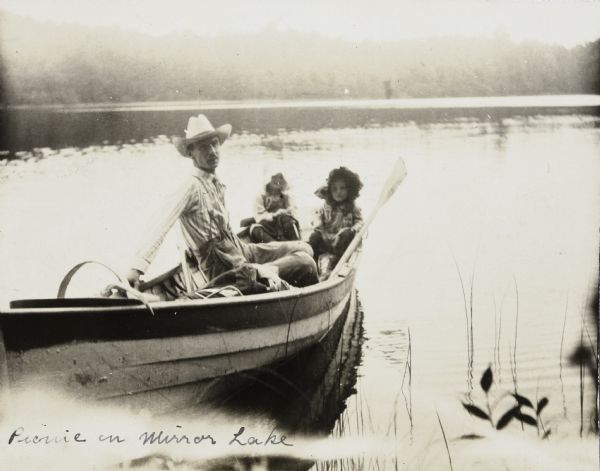 View from shoreline towards W.H. Young and two girls, all looking towards the camera, are sitting in a rowboat which is resting near the shore of Mirror Lake on a misty day. Caption reads: "Picnic on Mirror Lake." 