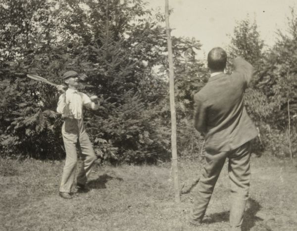 W.H. Young, holding a lawn tennis racquet, is playing tetherball against a man who has his back to the camera. A central wooden tetherball pole divides the scene. Trees are in the background. Page heading reads: "Tetherball."