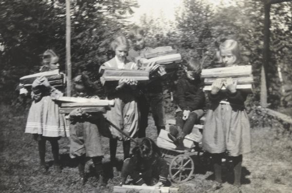 A group portrait of children carrying newly split wood in their arms. Alfred Holt, wearing glasses, is sitting on a pile of wood in a small wagon. Donald Holt, wearing bloomers, is second from the left. There are trees in the background. 