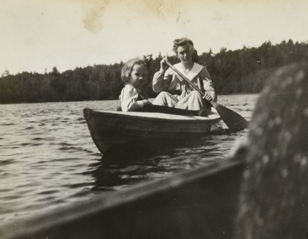 Harriet McClure is paddling a canoe on Archibald Lake, and Eleanor Holt is riding along. Both girls are looking at the camera. The side of a boat is in the foreground, and a tree-lined shoreline is in the background. Caption reads: "Harriet McClure and Eleanor canoeing." 