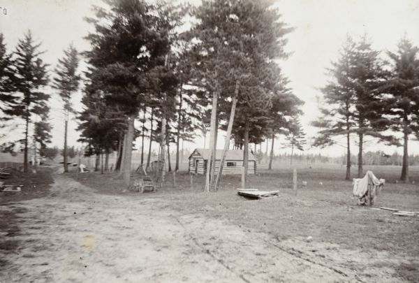 View down dirt road leading to the left, with a log cabin to the right among trees. There is a farm implement near the road. Caption reads: "At the Ranch."   