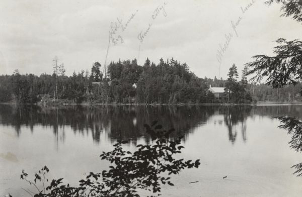 View from shoreline across Archibald Lake, looking north toward the Island. There is a view, from left to right, of three houses: Island Lodge, Green Cottage and the Wheeler Frame House, also known as The Ark. Hidden among the trees, only the Island Lodge roof can be seen. Green Cottage is barely visible. The Ark cottage, built close to the shoreline, can be seen among trees.