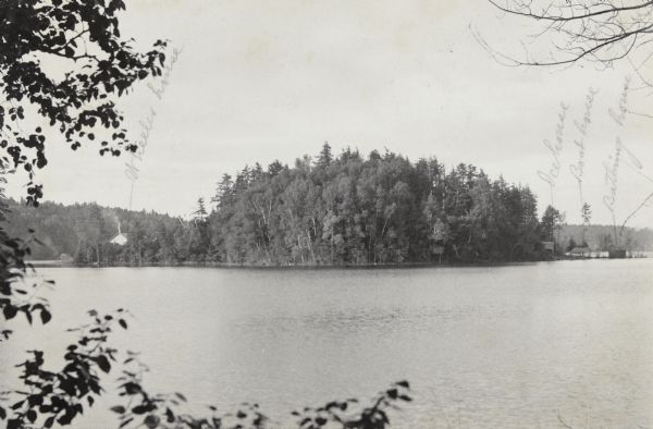 Elevated view from shoreline towards the tree-lined shore of the Island on Archibald Lake, and four buildings. From left to right the buildings are: Anna Wheeler's house, also known as The Ark; the ice house, the boathouse for the steam launch, and the bathing house. Smoke is rising from the chimney on The Ark cottage. 

