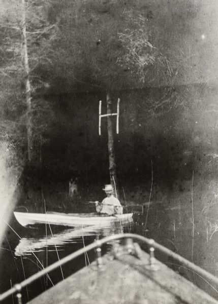 In the foreground is the bow of the Islander steam launch. George Holt, sitting in a clinker-built duck boat just beyond the launch, is holding the paddle with two hands. Above him, nailed to the tree, is hanging the "H," which was made of birch branches by Lucy Holt and Juliet Rumsey Stroh. The "H" stands for Hermit. In the background is the tree-lined shoreline of Hermit Island. Caption reads: "George Holt." 