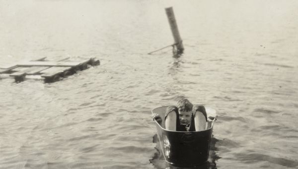 Squinting at the camera, Alfred Holt is lying in a metal tub, which is floating on Archibald Lake. His knees are bent and his hands are holding the sides of the tub. In the background is a wooden raft floating on the lake with a rope attached to a mooring post. Caption reads: "Alfred in Tub."