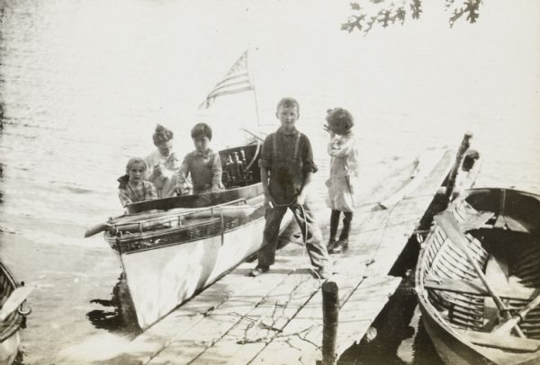 Elevated view from shoreline of Archibald Lake towards Alfred Holt standing on the pier holding the rope with both hands to hold the <i>Islander</i> steam launch, next to the pier on the left. A girl, possibly Eleanor Holt, is standing behind Alfred Holt and is rubbing her eye and looking down. A girl with a bow in her hair, possibly Jeannette Holt, is sitting in the front of the launch and looking at the camera. A boy is standing next to her, and a woman and baby are sitting in the back of the boat. A nautical flag is flying from the stern of the steam launch. A rowboat is docked on the right. A portion of another rowboat is on the lower left. 