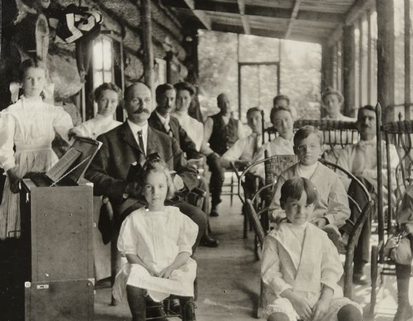 Sunday church service is being held on the screened porch at Island Lodge on Archibald Lake. Left to right, front row: Eleanor Holt, Donald Holt. Second row: Jeannette Holt (standing behind the pump organ), Arthur Dana Wheeler (sitting), and Alfred Holt (wearing glasses). Third row, behind Alfred Holt: Juliet Rumsey Stroh and Grant Stroh (sitting on wicker couch). Caption reads: "Church service."

