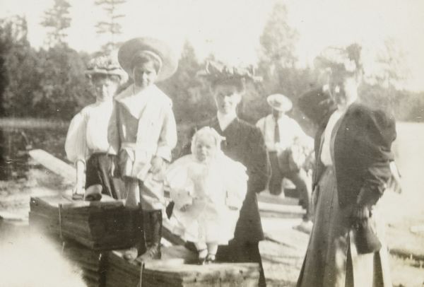 Baby Lillian Wheeler, wearing a dress, is standing on a pile of lumber with support from her mother Anna (Holt) Wheeler. Lillian's brother Gordon Wheeler, wearing a wide-brimmed hat, is also standing on the lumber, to the left of his sister with Ellen Holt behind him. Minnie May Rumsey is standing on the far right. In the background, Arthur Wheeler is getting out of the boat. Archibald Lake and the tree-lined shore are in the background. 

