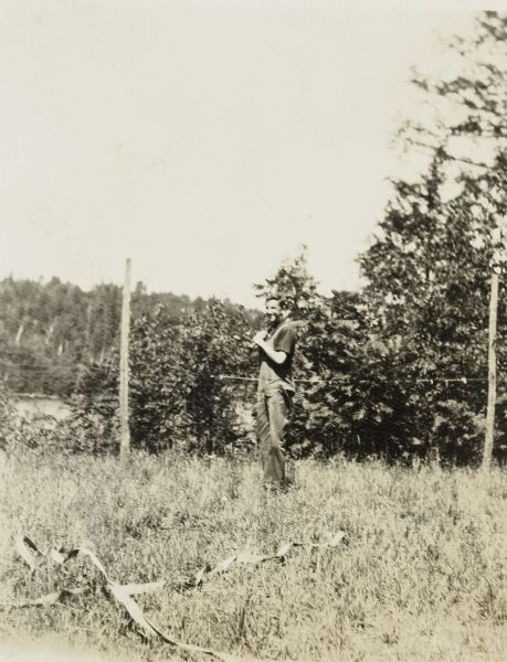 View across grass towards James McClure holding a baseball bat on his shoulder. Trees and bushes are in the background. Caption reads: "A baseball fan." 