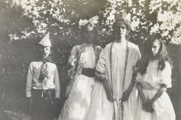 Four actors in costume are posing for a photograph with trees in the background. On the left is Alfred Holt wearing a hat and tie. To the right of Alfred is a girl wearing a big bow in her hair, then a girl wearing a crown on her head. On the far right is a girl with a headband with a small bow. Caption reads: "The Play — Hermit Island — Wed Aug 19 — Open Air Players."