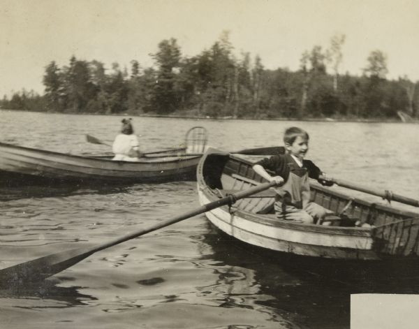 With her back to the camera, Eleanor Holt is rowing a boat on Archibald Lake. In the foreground, Donald Holt is rowing another boat. The tree-lined shoreline is in the background. Caption reads: "Donald, Eleanor."
