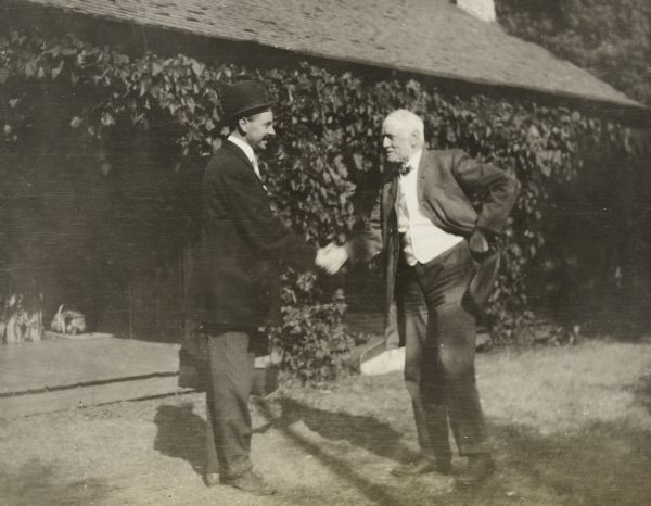 Seen in profile, Ralph Flanders and I.P. Rumsey are shaking hands and smiling at each other while standing in front of the Island Lodge porch. Ralph Flanders is wearing a bowler hat and is carrying a small case in his left hand. I.P. Rumsey is wearing a suit and bow tie, and is leaning slightly forward. Caption reads: "Till we meet again." Names written below are Ralph Flanders and I.P. Rumsey.