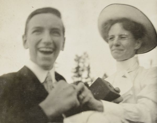 Close-up of Archibald McClure smiling. He is holding a box camera in his hands.  Next to him on the right is Lucy Holt, who is wearing a hat and eyeglasses, and is looking at Archibald McClure. They are holding hands. Caption reads: "Arch's farewell smile!" 