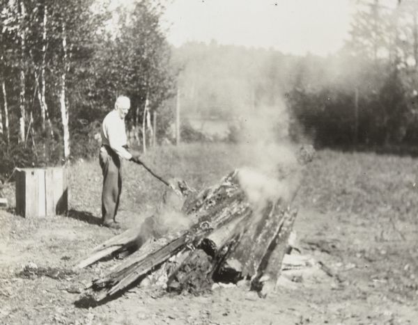 Wearing gloves, I.P. Rumsey is holding a stick and tending a smoking fire outdoors in a clearing on the Island. Archibald Lake is in the background.  