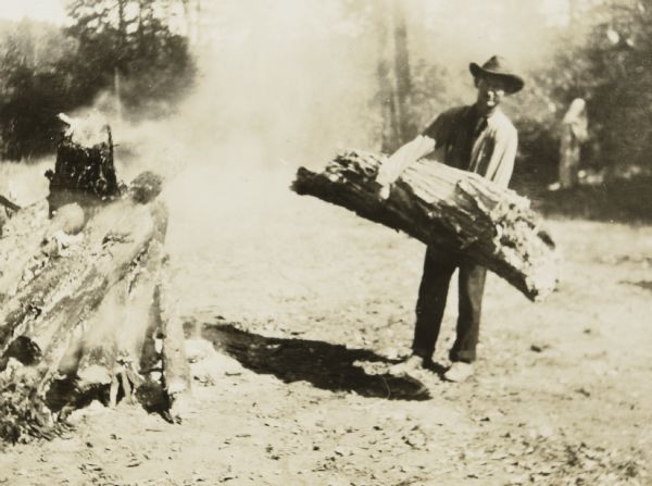 Looking at the camera, Albert Rhode, possibly a lumberjack, is standing and holding a big, heavy log. He is wearing a long sleeved shirt, pants, and a hat. A fire pile, with smoking rising from it, is on the left. Caption reads: "Albert Rhode." 