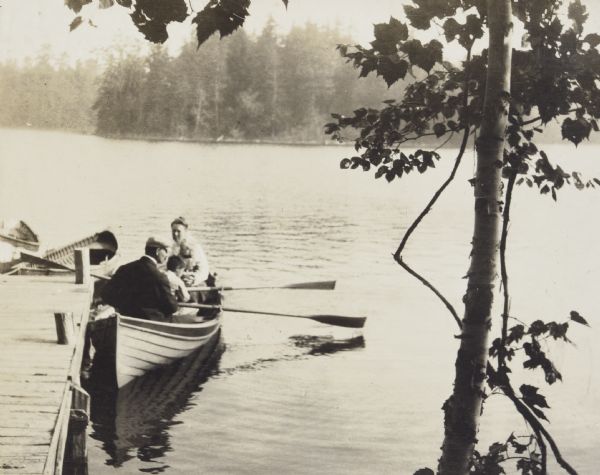 View from the Island shoreline towards Anna Holt Wheeler and husband Arthur Dana Wheeler, with children Gordon and Lillian, all sitting in a rowboat at the Ark dock, with Hermit Island in the background. Arthur and Gordon are holding oars. Lillian is sitting in her mother's lap. Two empty boats are docked to the end of the pier.  There is a tree on the right, in the foreground. Caption reads: "The Wheeler Family — Their Pier."


