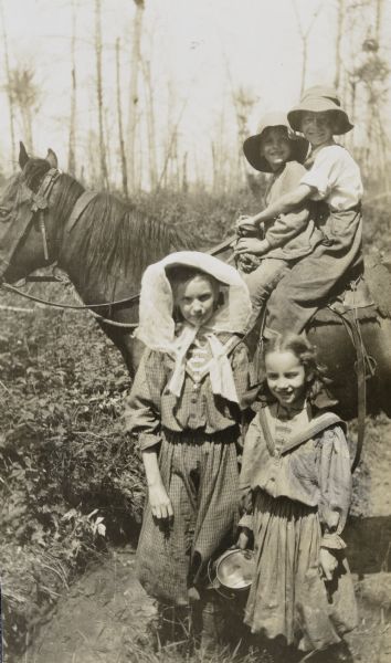 Brothers Alfred Holt and Donald Holt are on horseback together, with Donald sitting at the front of the saddle. On the left in front is Jeannette Holt, wearing a head scarf. She is standing next to her younger sister, Eleanor Holt, who is carrying a berry pail. Caption reads: "Holt children."