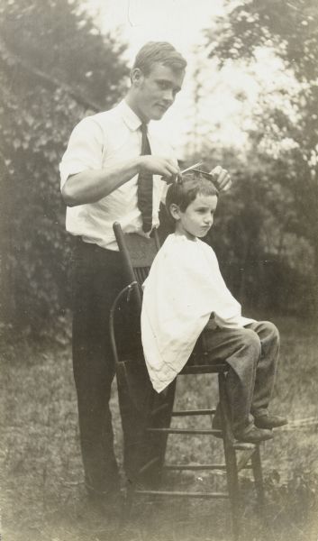 Donald Holt, who is seven or eight years old, is sitting in a chair outdoors while Joe Rumsey is cutting his hair. Caption reads: "Joe Rumsey and Donald."