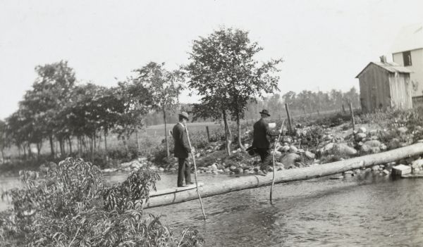 View from shoreline towards W.A. Holt and Mr. McDonald, dressed in suits, walking across a fallen tree trunk which serves as a bridge over a river. They are using walking sticks. Caption reads: "At the farm — WAH and Mr. McDonald."