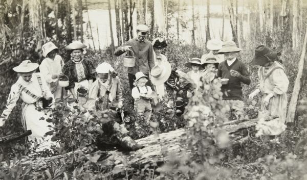 A group of twelve people is standing in the woods holding berry pails. Three generations are represented. Grant Stroh is in the center with a child in front of him. Alfred Holt, wearing glasses, is examining a berry on the far right. The rest of the group are seven women and a couple of children. Archibald Lake is in the background. 