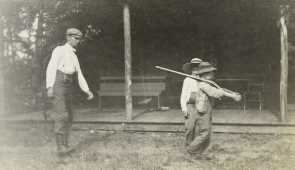 One boy, either Donald Holt or Gordon Wheeler, is marching and carrying a shotgun over his shoulder. The other boy, either Donald Holt or Gordon Wheeler, is standing in back of him watching. A man on the left, wearing a cap, is walking several steps behind the boy with the gun. Island Lodge porch is in the background. 

