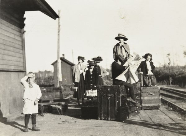 A group of women and children are waiting at the Lakewood Train Station. A young boy, with his hand shading his eyes, is standing next to the train station building on the left. Two women and a girl are waiting on the left. A boy, wearing a hat, is sitting on a large trunk and reading a big newspaper. Behind the boy, a person, wearing a coat, is standing on a trunk. Another girl is leaning on a trunk next to the railroad tracks on the right. Caption reads: "At the Lakewood Station 1909."                                                                                                                                                                       