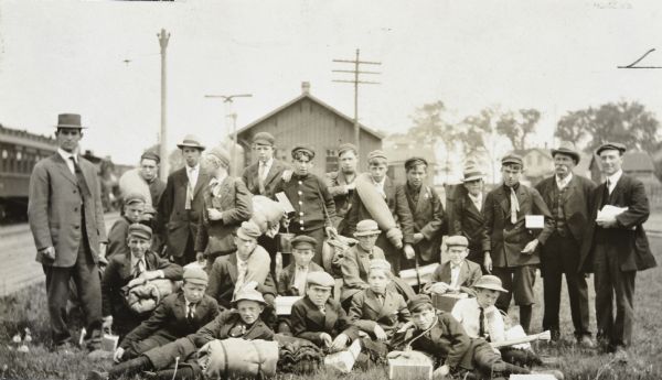 Members of the Boys' Brigade are posing for a group picture at the train station. Some boys are holding bedrolls. Three men, one on the left and two on the right, are standing with the boys. In the background is a railroad car parked on the tracks, the train station, an electrical pole, and a light pole. Caption reads: "Brigade Boys June 13th."