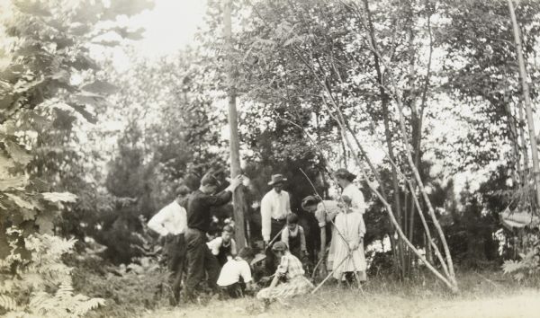 With shovels in hand, a man, woman, and boy are digging a hole in the ground for a flag pole.  One young man is holding the pole in place with two hands. A small group of children and adults is watching the pole raising. Archibald Lake and its tree-lined shore are in the background. 