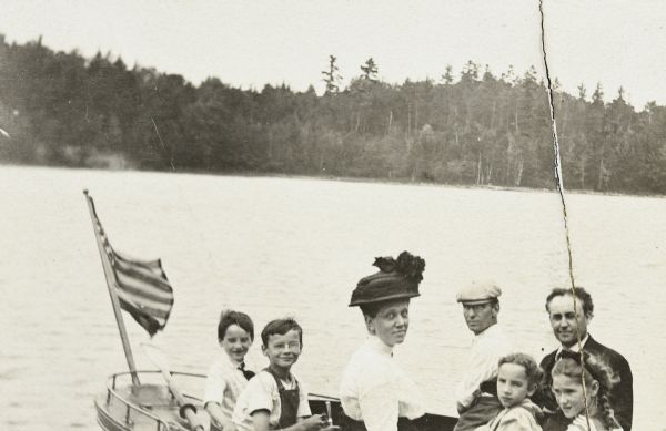Mr. and Mrs. Ramsey of Oconto are taking a ride in the <i>Islander</i> steam launch on Archibald Lake. Mrs. Ramsey is wearing a hat. Mr. Ramsey is wearing a suit. Grant Stroh is sitting to the left of Mr. Ramsey. The names of the children from left to right are: Donald Holt, Alfred Holt, Eleanor Holt and Jeannette Holt. The nautical flag is flying from the stern of the boat. 
