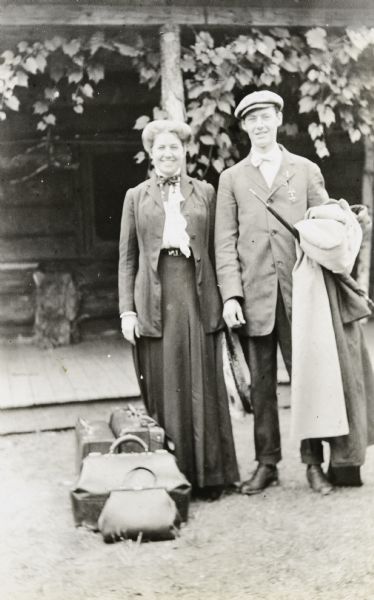 Edna and Wallace Rumsey, both smiling, are standing for a portrait in front of the Island Lodge porch. Wallace is holding a coat and umbrella. Their luggage is on the ground next to Edna. Caption reads: "Edna and Wallace Rumsey."