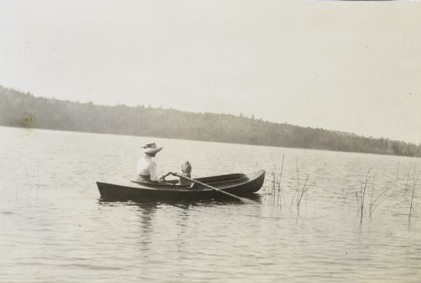 Lillian Wheeler is rowing a boat on Archibald Lake. She is looking at the oar. A woman, sitting in the rowboat and wearing a sun hat, is possibly Lillian's mother Anna Holt Wheeler. She is watching Lillian. The forested shoreline is in the background. 


